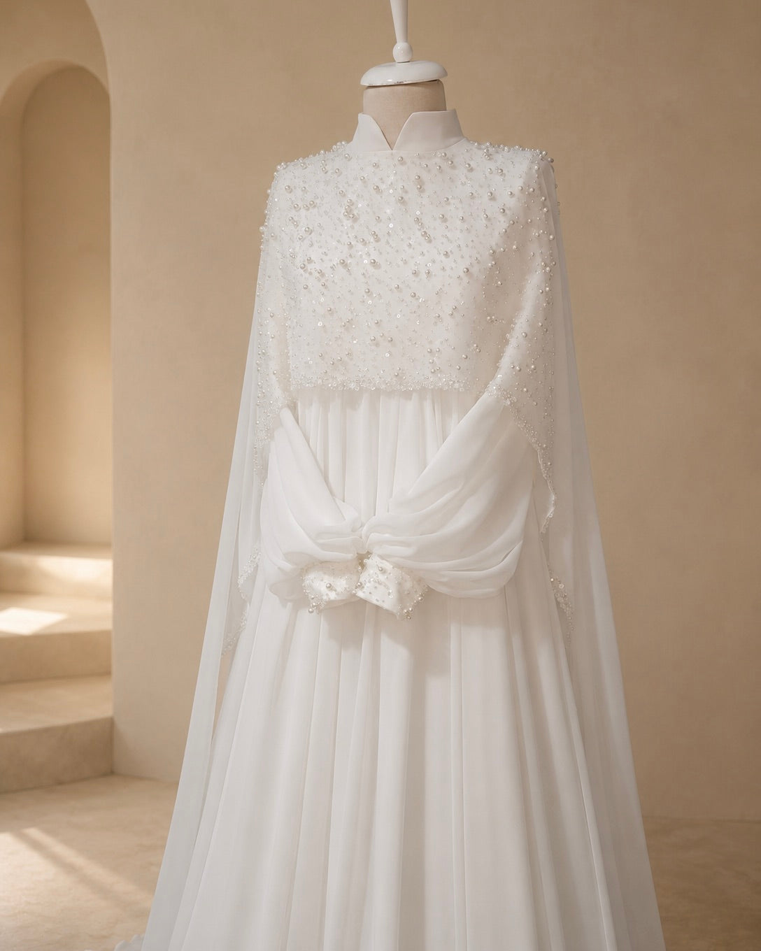A white bridal gown displayed on a mannequin in a softly lit beige interior. The dress features a high neckline, a heavily embellished beaded bodice, sheer draped cap sleeves, and a gathered waist with a bow detail.