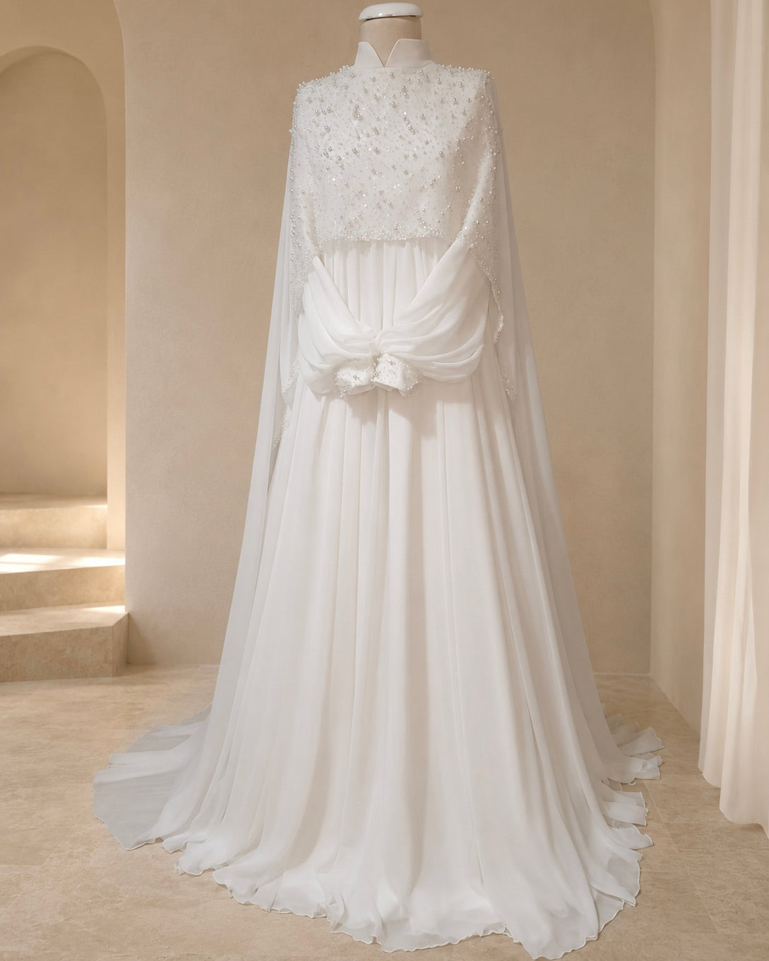 A white bridal gown displayed on a mannequin in a softly lit indoor boutique setting. The dress features a high neckline, a sheer embroidered bodice, a draped cape-like overlay, a gathered waist with a bow detail, and a flowing skirt with a train.
