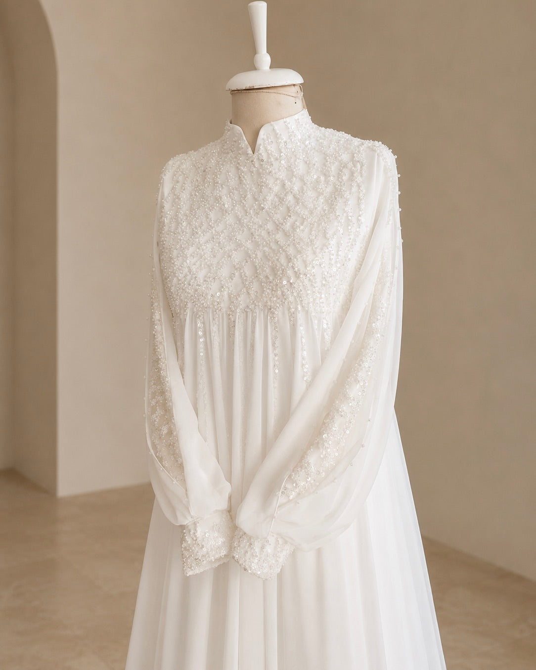 A white long-sleeve dress with intricate lace and embroidery on the bodice and cuffs, displayed on a cream-colored mannequin in a softly lit neutral interior.